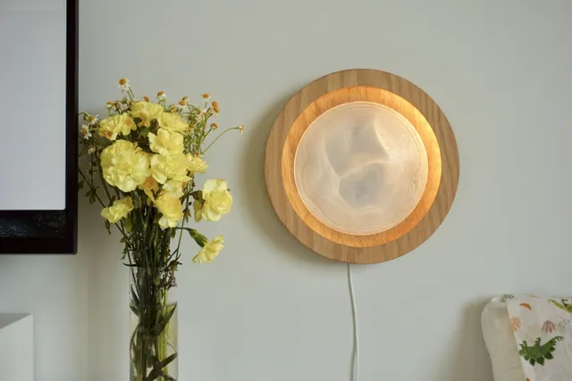 Round wooden Dune lamp glowing on a wall beside a vase of yellow flowers