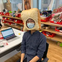 Jason Gao wearing a toast-shaped hat in a workshop filled with computers, tools, and electronics