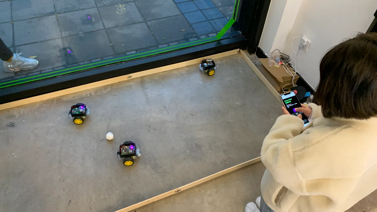 three robot on the ground moving a ball, a person next to using a mobile phone controlling on of the robot