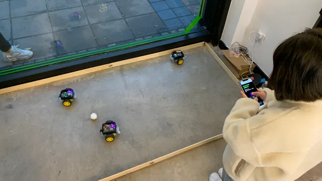 Person using a phone to control three small robot cars around a ball inside an enclosed arena