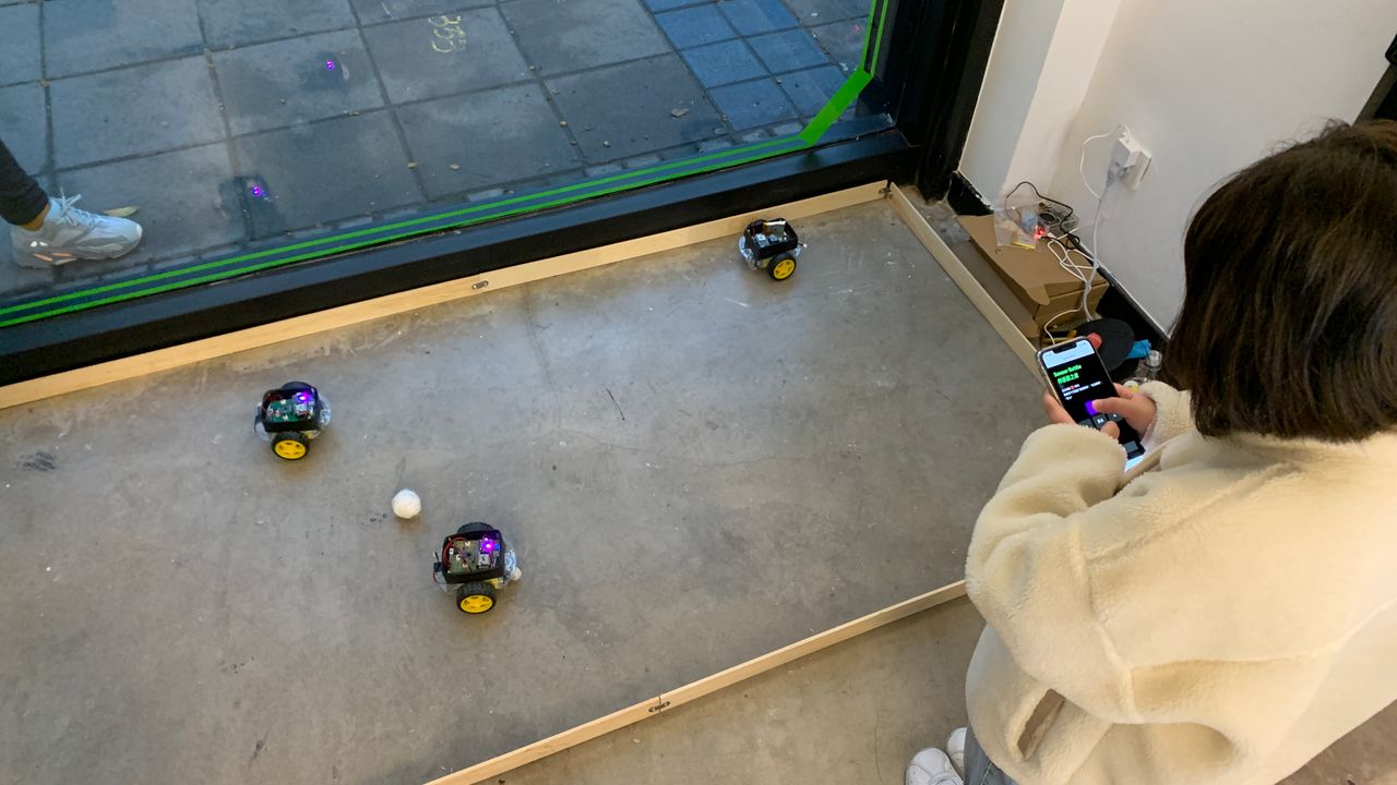 three robot on the ground moving a ball, a person next to using a mobile phone controlling on of the robot